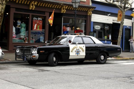 Black and white police car with 'Highway Patrol' on the door, parked on street in front of shops