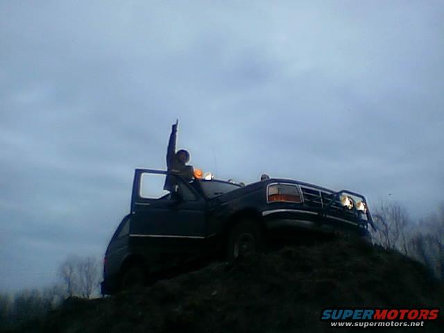 Dark blue Ford Bronco with a person on top, parked on a dirt mound under a cloudy sky