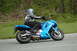 2007 Kawasaki Ninja R - A rider on a blue motorcycle cruises down a road, with trees and foliage in the background. The rider's jacket displays the Kawasaki logo.