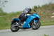 2007 Kawasaki Ninja R - A rider on a blue Kawasaki Ninja motorcycle cruises down a paved road with grassy hills in the background.