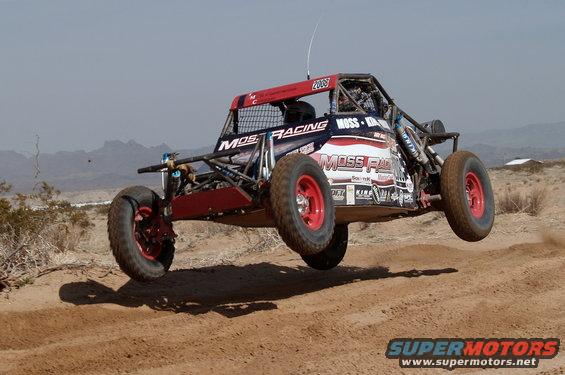 Off-road buggy with red roll cage and wheels airborne over dirt track. "MOSS RACING" text visible