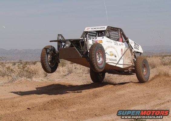 White off-road buggy with number 2022 airborne over a dirt track, desert landscape