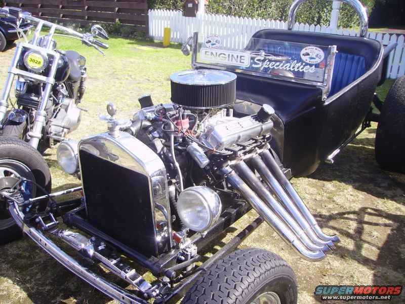 Hot rod with exposed V8 engine, chrome grille, and blue interior next to a motorcycle