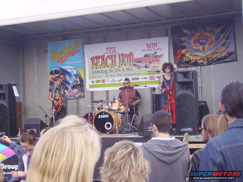 Band performing on a stage with banners behind them. A drummer, guitarist, and bassist are visible