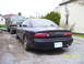 1997 Mazda MX-6 - A black coupe is parked in a gravel driveway next to a pickup truck, with houses in the background.