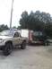 1982 Ford Bronco - A Toyota Tacoma tows a trailer carrying a disassembled vehicle body, with a utility building in the background.