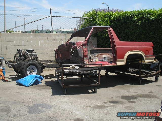 Disassembled red and tan Ford Bronco body and chassis on carts, engine separate, outdoors