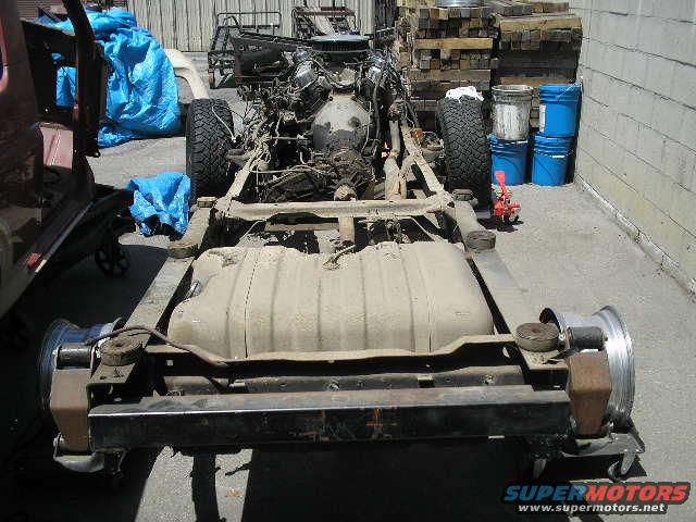 Vehicle chassis with engine and fuel tank, viewed from the rear. Partially disassembled, with blue tarps and stacked wood in background
