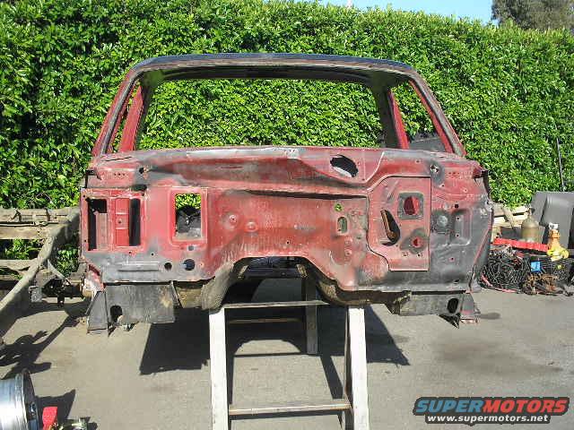 car body shell on stand, red paint stripped, green hedge behind