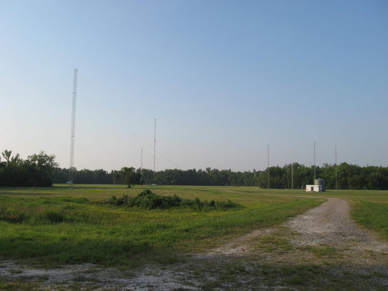 Wide shot of a field with several tall radio towers and a small white building under a clear blue sky
