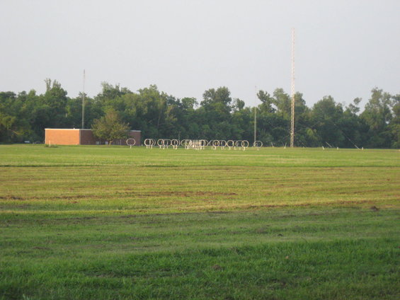 Wide view of a grassy field with a brick building, a row of circular antennas, and a tall radio tower in the background