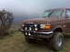 1990 Ford Bronco - A brown Ford truck with a brush guard and large off-road tires sits in a grassy, overcast landscape.