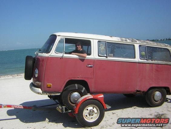 Red and white VW bus on a trailer at the beach, person in driver's seat