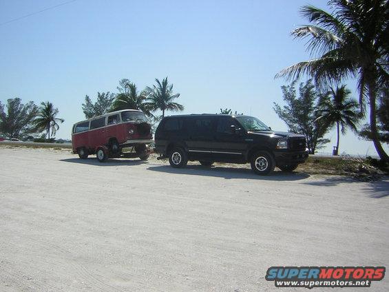 Red and white Volkswagen Type 2 van being towed by a black SUV on a sandy road