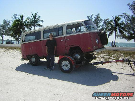 red and white van on a U-Haul trailer, person standing next to it, palm trees and ocean in background
