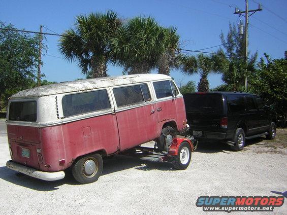 Red and white Volkswagen bus on a trailer, seen from the rear quarter, with a black SUV behind it