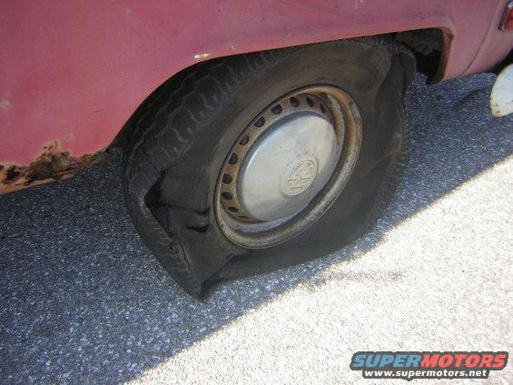Flat tire on the rear passenger side of a red vehicle with visible rust on the fender