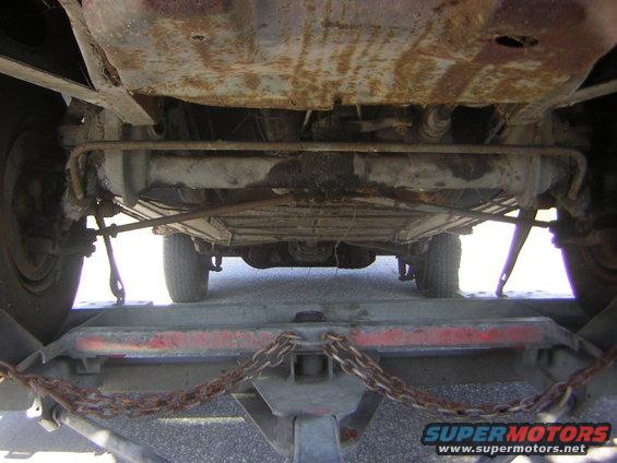 Underside view of a vehicle being transported on a trailer, showing rusty undercarriage and suspension components