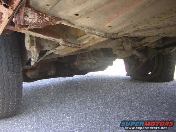 Underside view of a rusty vehicle, showing the drivetrain and suspension components. One tire is visible on the left