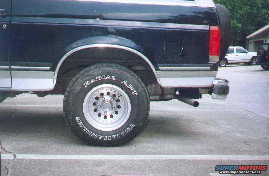 1990 Ford Bronco rear side view showing wheel, fender, and part of the tailgate. Another white SUV is parked in the background