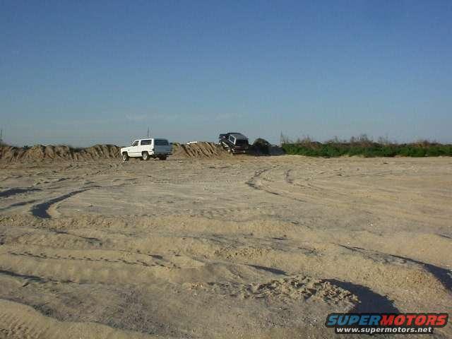 White SUV parked on sand, black SUV on a dirt mound under a clear blue sky