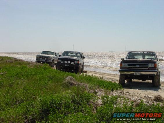 1990 Ford Bronco and two other Ford trucks parked on a sandy beach near the water