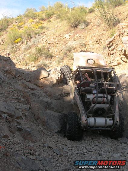 Custom off-road buggy with roll cage and large tires navigating a steep, rocky hill. Watermark reads SUPERMOTORS.NET