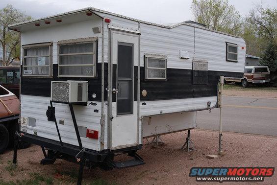 Slide-in truck camper with black stripe, parked outside. Another truck is visible in the background