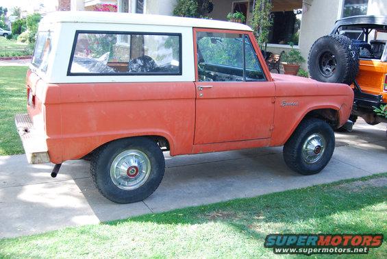 Orange and white Ford Bronco, side view, weathered paint, chrome hubcaps, parked on concrete driveway