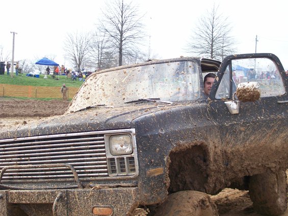 Mud-covered truck, front 3/4 view, with driver visible. Grille and headlights are prominent