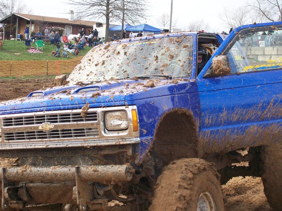 Muddy blue pickup truck with open driver's door, covered in mud splatters. Grille has horizontal bars and a Chevrolet emblem
