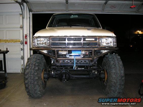 White Toyota truck front view with large off-road tires, winch, and custom bumper. Grille has "TOYOTA" text