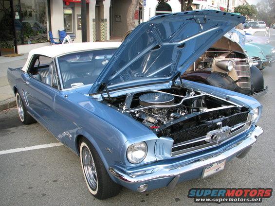 Blue convertible with white top, hood open, showing chrome engine details. Whitewall tires. A 1965 Ford Mustang
