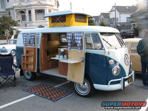Blue and white VW camper van with pop-top, open doors, and interior setup. Another car is parked nearby