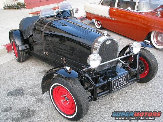 Black vintage roadster with red wheels and a sign that says 'ASK ME IF I REALLY GIVE A CHIT'. Another car is parked behind it