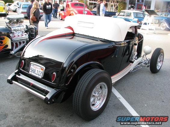 Black hot rod convertible with white top, chrome bumper, and California plate 5HVL112. Other cars and people in background
