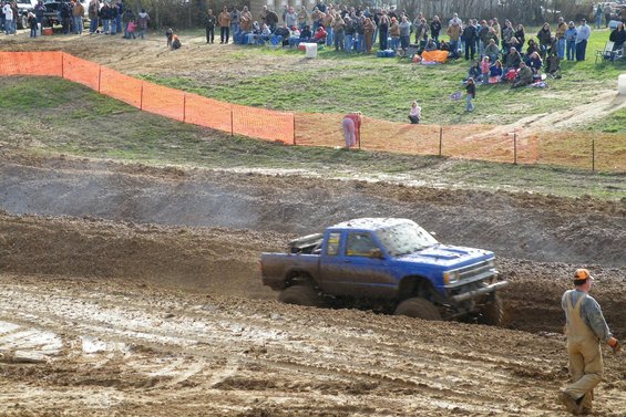 Mud-splattered blue pickup truck with lifted suspension driving through a mud pit