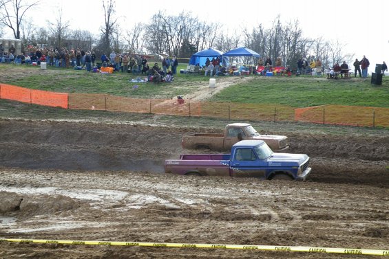 Blue and purple pickup truck stuck in mud pit with spectators in background