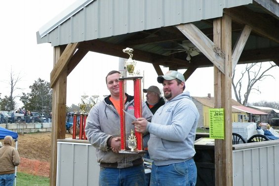 Two men holding a large trophy under a wooden pavilion, with other trophies and vehicles in the background