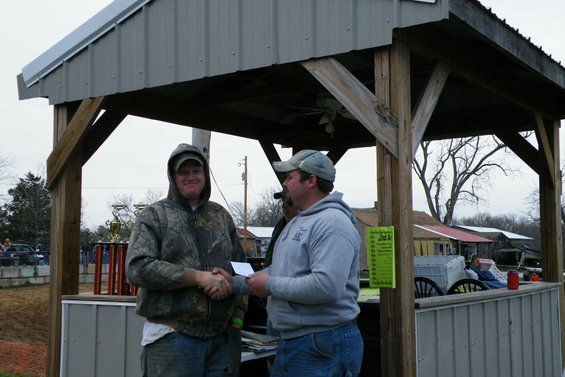 Man in camo jacket shakes hand with man in gray hoodie under a covered structure. Trophy visible