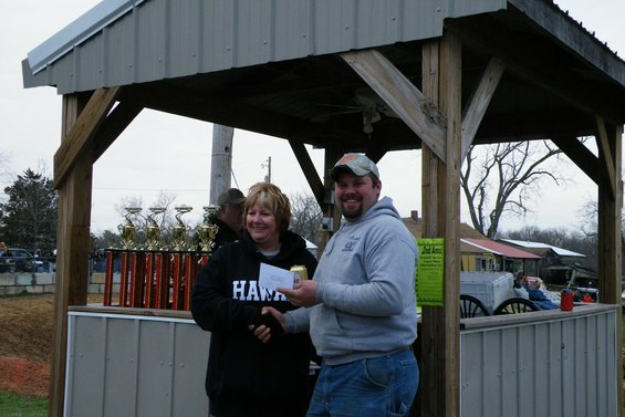 Man in grey hoodie shakes hands with woman in black hoodie, trophies behind them
