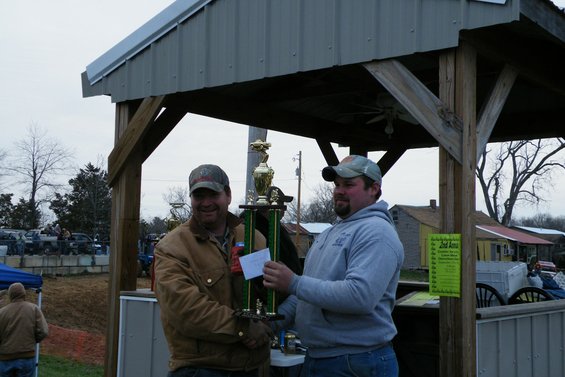 Two men holding a trophy at an outdoor event with vehicles in the background