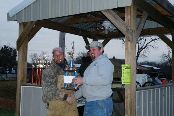 Man in camouflage jacket and apron shakes hand of man in gray hoodie, both holding trophies. Yellow sign on post behind them
