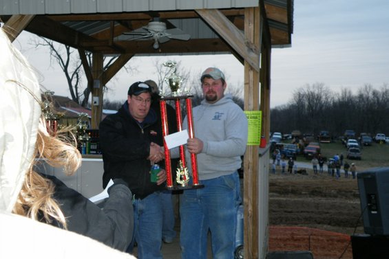 Two men holding a large trophy, one shaking hands, with a crowd and parked vehicles in the background