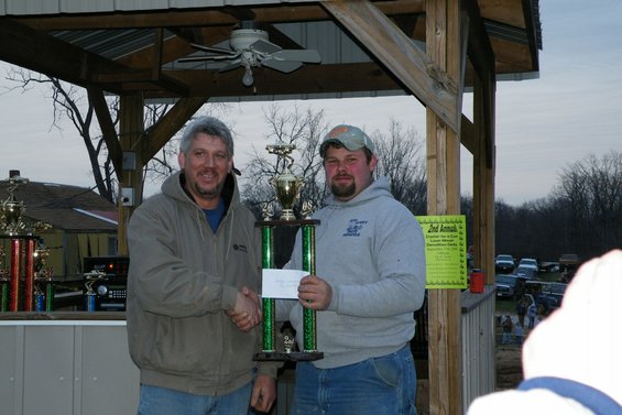Two men shake hands while holding a trophy, with parked cars in the background