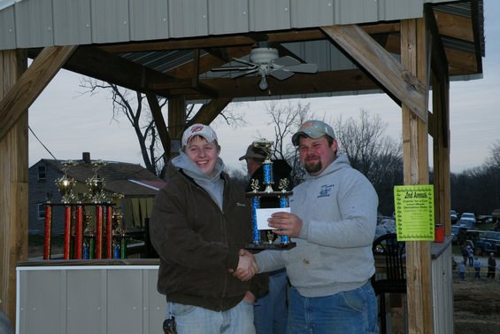 Two men shake hands under a covered structure, one holding a trophy. Several other trophies are visible on a shelf. Vehicles are parked in the background