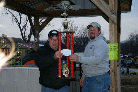 two men holding a large trophy, one handing the other a piece of paper. A sign is visible behind them