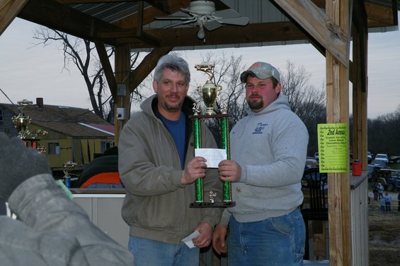 Two men hold a trophy with a check. A yellow sign is on a wooden post