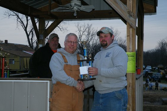 Two men shake hands while receiving a trophy, with vehicles visible in the background