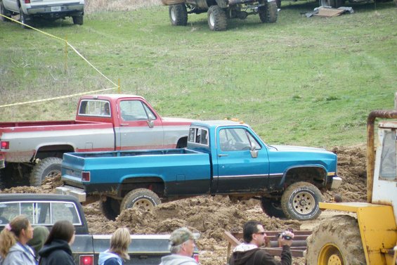 Blue pickup truck stuck in mud, side view. Red pickup truck behind it